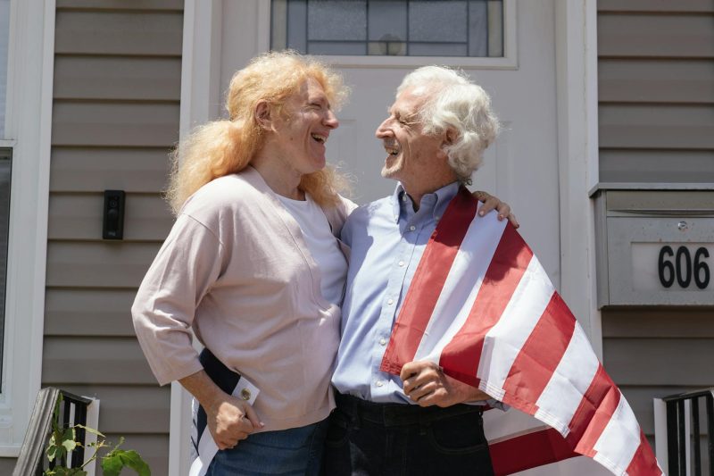 Happy senior couple standing outdoors, smiling together with an American flag waving in the background.