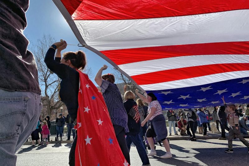 Celebrators raising hands beneath a large American flag at a lively parade in Denver, CO.