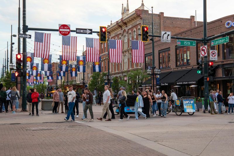 Crowd of people crossing a vibrant downtown Denver intersection, surrounded by colorful flags.
