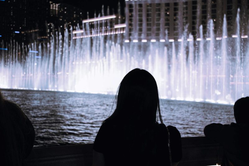 Silhouette of a person watching a colorful illuminated fountain show in a bustling urban setting at night.